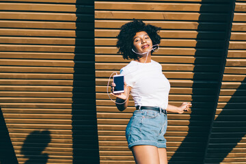 Cheerful happy african american meloman excited with good music sound in earphones jumping feeling carefree, emotional dark skinned hipster girl dancing to playlist songs on weekends in park