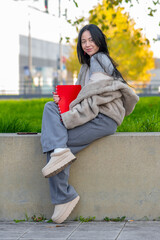 A young woman of 20-25 years of oriental appearance sits on a low wall with a folder in her hands, enjoying a sunny day in a city park, 