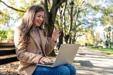 Young businesswoman on video call using laptop outdoors in city park, engaging in remote work. Professional woman online connecting with employees and clients for global business.