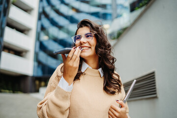 Young businesswoman sending voice message in front of modern business building	