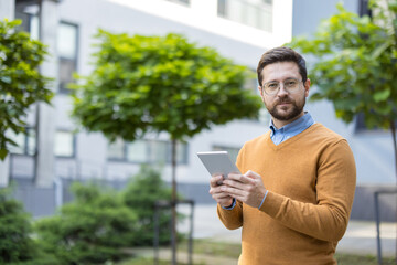 Portrait of a young man standing on the street, holding a tablet in his hands, looking seriously and confidently into the camera