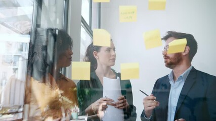 Video of successful business team writing on stickers at office glass board while discussing together in the coworking space.