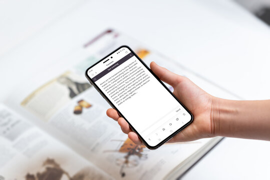 Hand holding smartphone scanning text in book, representing technology use for extracting knowledge, information, and details from printed materials