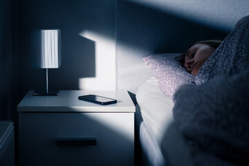 Phone next to bed while sleeping. Mobile on table and nightstand at night. Woman and shut down cellphone at bedside. Light from window. Young person asleep in bedroom. Alarm clock in smartphone.