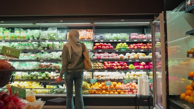 A female shopper walking in store and holding cart with fresh fruits. A young woman carries a shopping cart full of organic products and walks inside the store, choosing healthy products and enjoying
