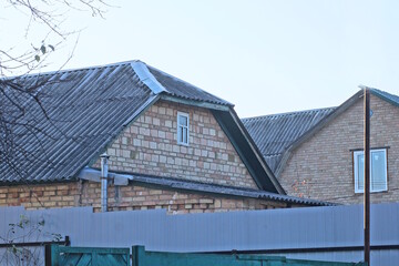 brown brick attic of a rural house with a small window under a gray slate roof behind a metal fence against the sky