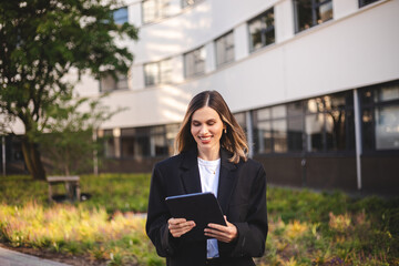 Smiling professional business woman bank manager, happy female executive or lady entrepreneur holding digital tablet pad standing near office at work on the street. Girl in suit read news, messages. 