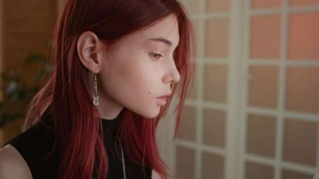 Tilt-up shot of teenage girl with red hair applying bronzer to eyes and nose, using cosmetic brush and looking in hand mirror while doing makeup in cozy living room