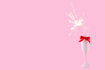 A champagne glass with a a red bow and sparkler on a pastel pink background. Celebration event concept. Holiday idea. Copy space.