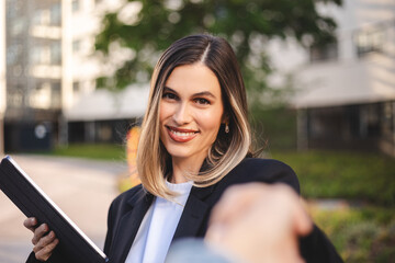 Confident businesswoman in blazer suit smiles, shakes hands with businessman outside modern city office. Holding tablet, negotiating successful partnership deal. Happy teamwork in urban setting.