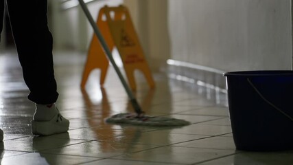 Close-up of a janitor mopping a tiled hallway floor with a bucket and caution sign visible, emphasizing workplace hygiene and safety. Concept of professional cleaning and maintenance.