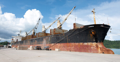 The rusty deck cranes on the large industrial cargo ship is loading many containers to its holds at the pier, Phuket, Thailand.