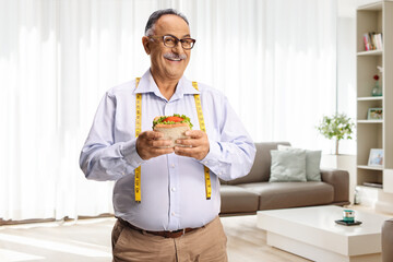 Hungry elderly man with a measure tape holding a sandwich at home