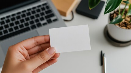 A hand displays a blank business card in a soft office environment featuring a laptop and a decorative plant. This setting suggests a professional context for networking or branding