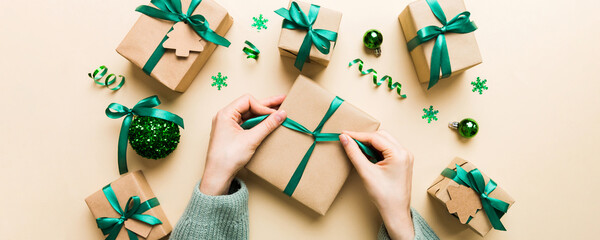 Woman is packing surprise gift for Christmas holidays, sitting at workplace with craft tools, top view. Woman wrapping Christmas gift at colored table