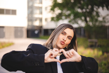 Happy attractive business blonde woman showing shape heart with hands, girl wear black jacket and white shirt standing near office in city. Love, romantic concept. Woman laughing and send blow kiss.