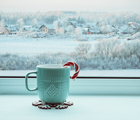 Winter background, cup with candy cane on windowsill and winter rural landscape outdoors. Cozy...