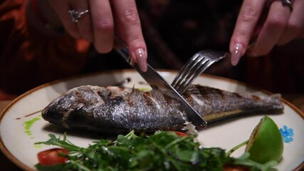 Close-up side view of woman cutting and removing skin from fried seabass (sea bass or branzino) fish lying on white plate on wooden table in restaurant. Soft focus. Beautiful and delicious food theme.
