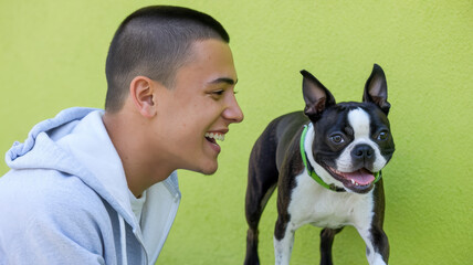 A joyful Hispanic teenage boy laughing with his playful Boston Terrier against a vibrant green background.