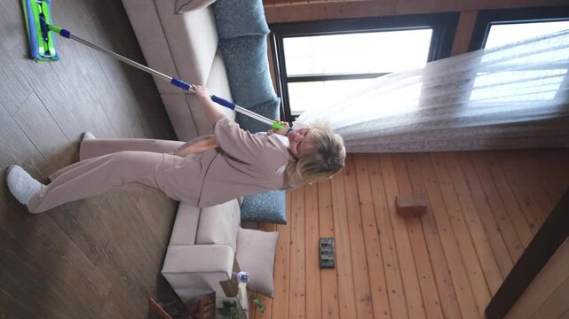 Happy young woman singing while washing floor at living room, using mop as microphone, having fun during house chores. Millennial lady enjoying domestic duties, gamifying cleaning routine