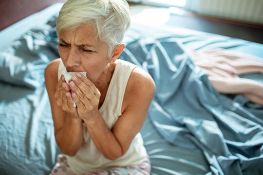 Senior woman with cold using tissue and nasal spray in bedroom