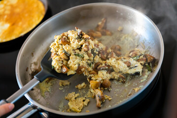 There is frying pan with scrambled eggs on stove. As flavor additive, pieces of champignons and green asparagus shoots were placed in dish.