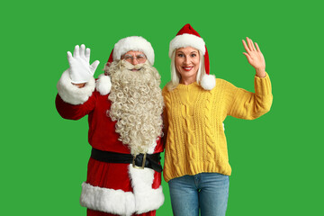 Santa Claus and beautiful mature woman in Christmas hats waving hands on green background