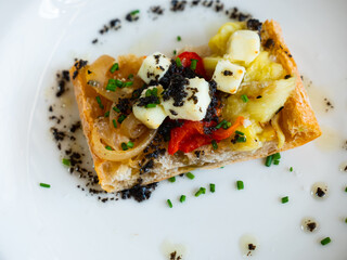 Plate contains sandwich made from piece of French baguette, stewed Escalibada vegetables, and cubes of goat cheese. Appetizer flavored with olive oil, served with glass of red wine.