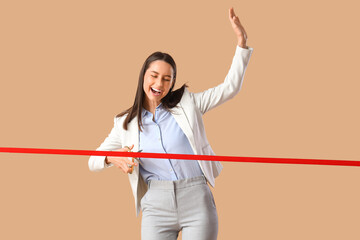 Happy young businesswoman cutting red ribbon on brown background