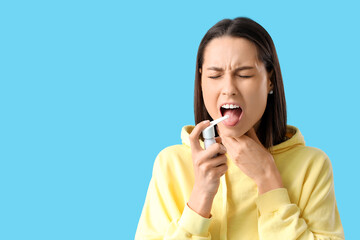 Young woman using spray for sore throat on blue background
