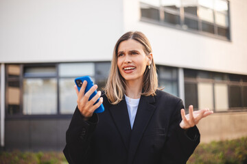 Angry young businesswoman in black jacket stands office outdoor records audio message, screams with...