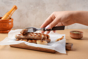 Woman holding fork and baking paper with tasty grilled sausages on beige table