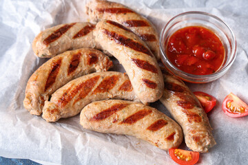 Baking paper with tasty grilled sausages, tomatoes and bowl of sauce on blue background, closeup