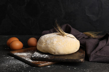 Wooden board with fresh raw dough, wheat and eggs on black background