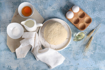 Plate with fresh raw dough, wheat, sugar, jug of milk and wooden holder of eggs on blue background