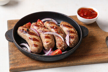 Frying pan with tasty grilled sausages, onion, tomatoes and bowl of sauce on white background
