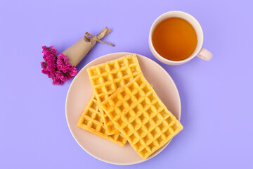 Delicious Belgian waffles in plate with cup of tea and flowers on lilac background. Top view