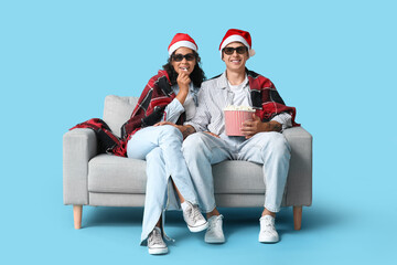 Beautiful young couple in Santa hats with bucket of popcorn watching movie and sitting on sofa against blue background. Christmas celebration