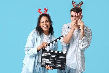 Beautiful young couple in reindeer horns with clapperboard and candy canes on blue background....