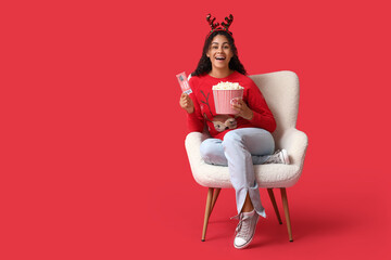 Beautiful young African-American woman with cinema ticket and bucket of popcorn sitting in armchair...