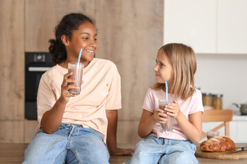Cute girls with glasses of sweet chocolate milk sitting on table in kitchen