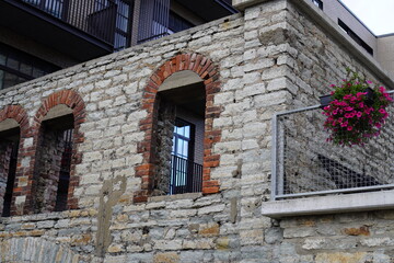 Historic stone wall with arched windows and hanging flowers near modern building in an urban setting during daylight hours. Kalamaja, Tallinn, Estonia, Europe