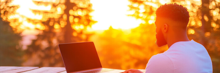 Man working on laptop with sunset in background.
