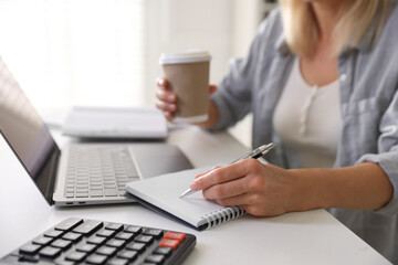 Budget planning. Woman with paper cup of coffee taking notes at table indoors, closeup