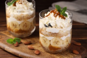 Tasty trifle dessert. Sponge cake, banana and whipped cream in glasses on wooden table, closeup