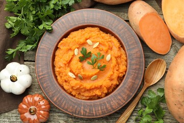 Tasty mashed sweet potato with pumpkin seeds in bowl, spoon, fresh vegetables and parsley on wooden table, flat lay