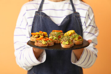 Young woman holding board with tasty vegan bruschettas on orange background