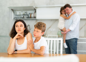 Offended mother sitting at the kitchen table being hugged by little son, her husband and daughter...