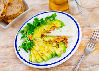 Appetizing pieces of cheese served with sliced avocado arugula and walnuts at table