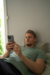 a young handsome man in a green t shirt lies in bed and stares at his phone